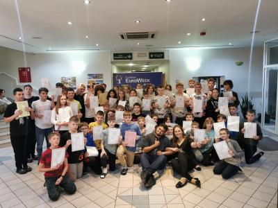 Grupowe zdjęcie wszystkich uczestników projektu wraz z trenerem i koordynatorką wyjazdu na tle plakatu EuroWeek. Uczestnicy trzymają w rękach certyfikaty uczestnictwa w obozie językowym.