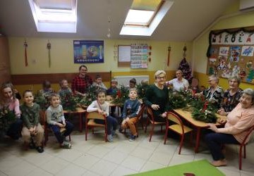 Dzieci z przedszkola wraz z nauczycialkami oraz zaproszonymi seniorami pozuja do zdjęcia. Na stołach stroiki świąteczne.