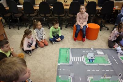 Uczniowie siedzą wokół makiety miasta uczestnicząc w zajęciach sztucznej inteligencji. Na makiecie postawione są budynki sklepów i urzędów.