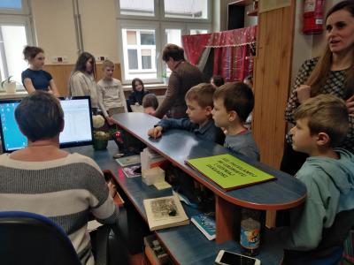 Pani bibliotekarka siedzi przed monitorem i wypożycza uczniom, którzy stoją przy ladzie biurka, wybrane przez nich książki.