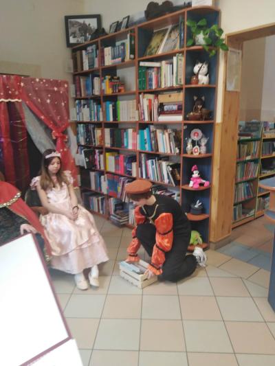 Na krześle siedzi uczennica przebrana za księżniczkę. Przed nią klęka uczennica przebrana za żaka i kładzie na podłodze skrzyneczkę z książkami.