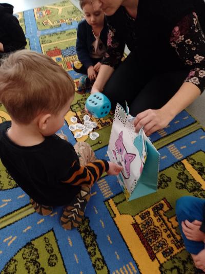 Chłopiec siedzi na dywanie i wrzuca myszki do buzi papierowego kotka naklejonego na torbę papierową. Nauczycielka trzyma kostkę do gry