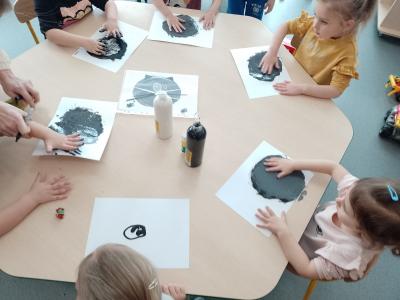 Dzieci siedzą przy stolikach i malują całą dłonią rozcierając farbę czarną a potem białą. Na stoliku stoi farba biała i czarna oraz zdjęcie gotowego kotka.
