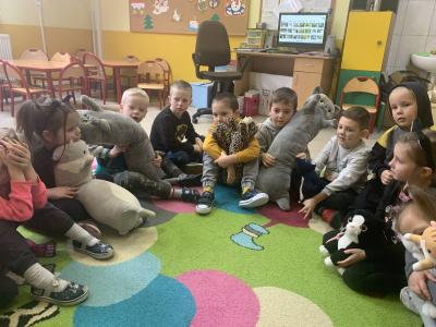 Dzieci siedzą w kole na dywanie w sali przedszkolnej i trzymają w dłoniach przyniesione z domów pluszaki kotów