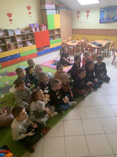 Dzieci siedzą w sali przedszkolnej na dywanie i oglądają wyświetlany na komputerze film edukacyjny o kotach.