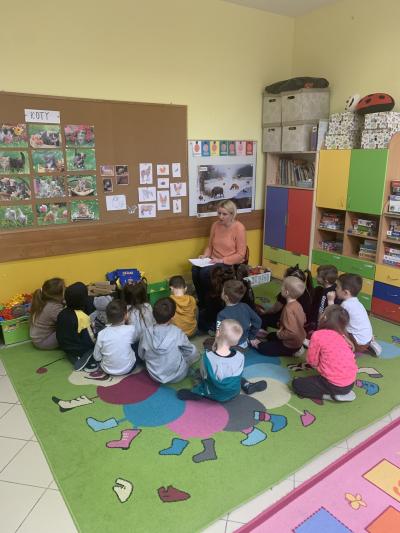 Dzieci w skupieniu siedzą na dywanie przed nauczycielem. Nauczyciel siedzi na małym krzesełku z kartką na kolanach i czyta bajkę. Na ścianie wisi tablica z obrazkami kotów