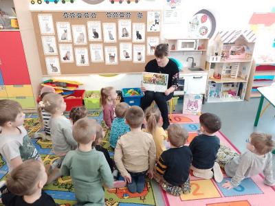 Na zdjęciu widać dzieci z grupy przedszkolnej „Krasnale”, które siedzą na kolorowym dywanie i patrzą w stronę tablicy. Nauczyciel siedzi na krzesełku i czyta dzieciom bajkę