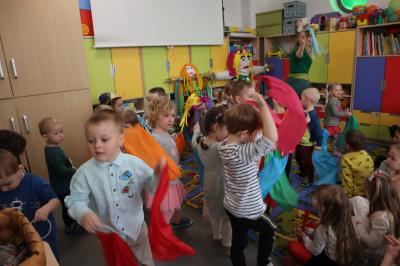 Nauczycielka przebrana za Pania Wiosnę tańczy z dziećmi, wszyscy machają kolorowymi szarfami.