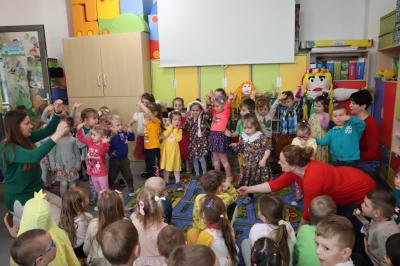 Dzieci z grupy Sówki pokazują ruchy do piosenki wiosennej, z lewej strony zdjecia nauczycielka unosi rece do góry, pokazuje ruchy.