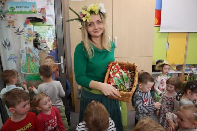 Nauczycielka przebrana za Panią Wiosnę uśiecha się, w rekach trzyma koszyk z cukierkami i nasionami owsa. Wokół stoją dzieci.