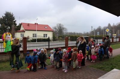 Dzieci z grup Sówki i Misie razem z paniami pozują do zdjęcia. Jedna z pań trzyma marzannę. Stoją przy szkole, za nimi ogrodzenie, w tle widać zaparkowane samochody.