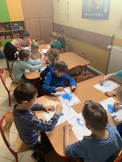 Dzieci siedzą przy stolikach w sali przedszkolnej i zamalowują niebieską farbą sylwety kropli.