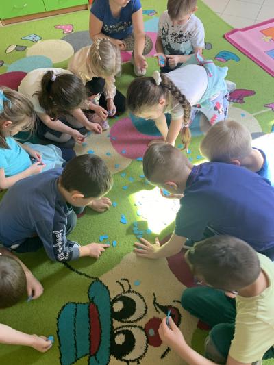 Dzieci zbierają rozsypane na dywanie w sali przedszkolnej małe, niebieskie, papierowe kropelki wg ilości podanej przez nauczyciela prowadzącego.