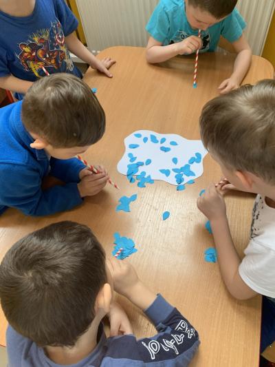Dzieci przenoszą za pomocą rurki do picia rozsypane na stoliku małe, niebieskie, wycięte z papieru kropelki na białą papierową chmurkę.