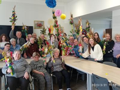 Wspólne zdjęcie seniorów wraz z wolontariuszami, którzy trzymają w dłoniach uniesionych do góry wykonane palmy wielkanocne.