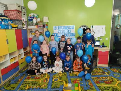 Wspólne zdjęcie dzieci z grupy Skrzaty wraz z wychowawczyniami. W tle tablica z dekoracją z okazji Światowego Dnia Świadomości Autyzmu. Dzieci trzymają w rękach niebieskie balony.