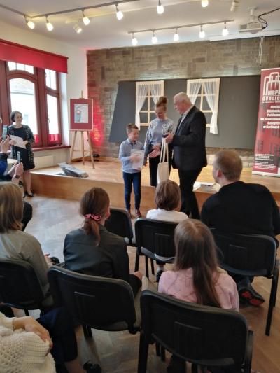 Na krzesłach siedzą uczestnicy konkursu wraz ze swoimi opiekunami. Na środku Sali stoi laureat konkursu oraz osoby, które wręczały mu nagrody, czyli pani Dyrektor Biblioteki w Gorlicach i przedstawiciel Starostwa.