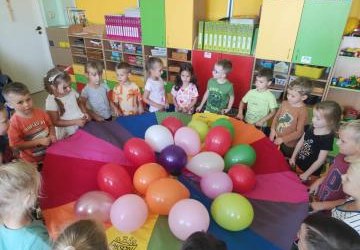 Dzieci trzymające kolorową hustę z balonami w środku.