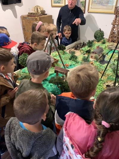 Dzieci zgromadzone są wokół makiety wykonanej przez młodzież przedstawiającej plan Skansenu. Przy ścianie stoi właściciel i pokazując dłonią objaśnia zgromadzone na makiecie miniaturowe eksponaty.