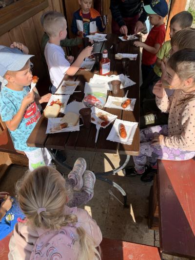 Dzieci siedzą  przy stole na zewnątrz budynku i jedzą kiełbaskę upieczoną na grilu z ketchupem, chlebem i herbatką w kubeczkach.