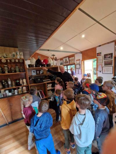 Dzieci zgromadzone są przed właścicielem Skansenu, który pokazuje zgromadzone na półce czapki górnicze z białymi, czarnymi i czerwonymi piórami. W tle widać kufle i medale górnicze, maszyny do pisania, książki o tematyce górniczej.