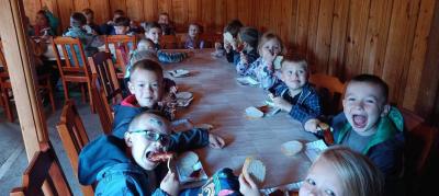 Dzieci siedzą przy stole wewnątrz drewnianego budynku i wesoło z apetytem spożywają upieczoną na grillu kiełbaskę wraz z chlebem i ketchupem położoną na papierowych tackach.