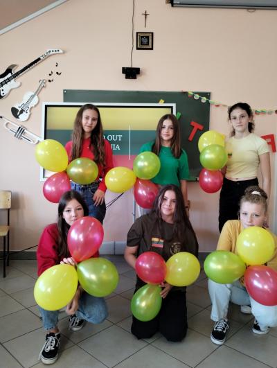 Sześć uczennic z balonami w kolorze żółtym, zielonym i czerwonym stoi na tle litewskiej flagi