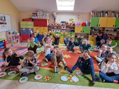 Dzieci siedzą na dywanie w sali przedszkolnej. Podnoszą do góry dłonie pokazując 10 palców jako wynik dodawania 5 czerwonych i 5 zielonych sylwet jabłek rozłożonych po równo na plastikowych białych tackach.