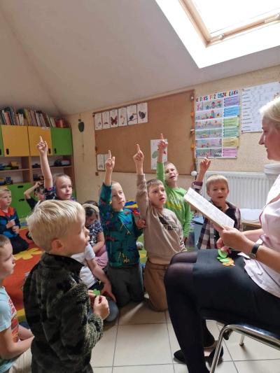Nauczyciel siedzi na krześle przez zgromadzonymi na dywanie dziećmi. Kilkoro dzieci podnosi palec do góry jako chęć udzielenia odpowiedzi na przeczytaną przez nauczyciela zagadkę o owocach i warzywach.