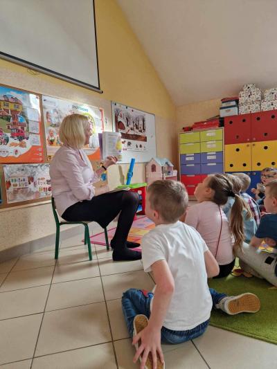 Nauczyciel siedzi na krześle w sali przedszkolnej i czyta dzieciom wiersz o owocach i warzywach Dzieci z uwagą słuchają tekstu i wpatrują się w obrazki w książce.