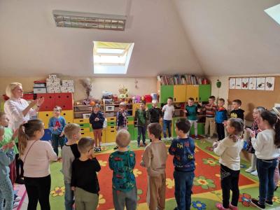 Dzieci stoją zgromadzone w kole na dywanie w sali przedszkolnej. Wśród nich jest nauczyciel, który śpiewa piosenkę na powitanie rozkładając dłonie do klaskania.
