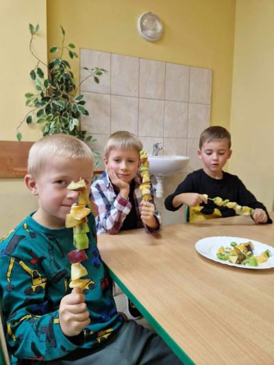 3 chłopców siedzi przy stoliku i trzyma w dłoniach zrobione szaszłyki z pokrojonych owoców.