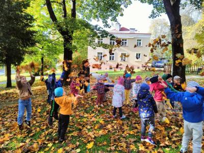 Grupa Sówki w parku. W tle widać budynek kina Farys w Bieczu. Dzieci są na trawie,  wokół nich liście kolorowe jesienne. podrzucają liście i skaczą, uśmiechają sie.