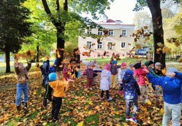 Grupa Sówki w parku. W tle widać budynek kina Farys w Bieczu. Dzieci są na trawie,  wokół nich liście kolorowe jesienne. podrzucają liście i skaczą, uśmiechają sie.