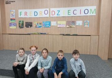 Pięciu uczniów siedzi na schodku w sali bibliotecznej. Nad  nimi wisi napis Fredro Dzieciom i prace uczniów.