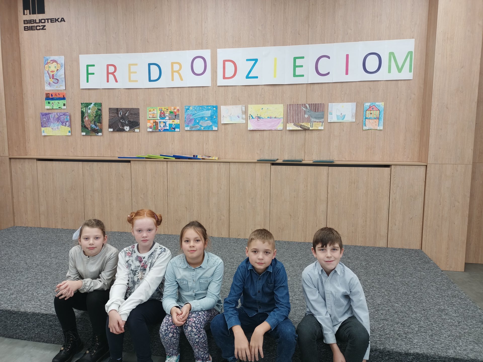 Pięciu uczniów siedzi na schodku w sali bibliotecznej. Nad  nimi wisi napis Fredro Dzieciom i prace uczniów.