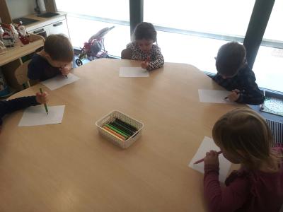 Dzieci siedzą przy stole i próbują narysować słoneczko ręką, którą na ogół się nie posługują.
