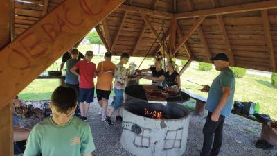 Uczestnicy ogniska gromadzą się pod wiatą ogniskową, odbierają kiełbaski, które spożywają. Rodzice pomagają w ognisku.