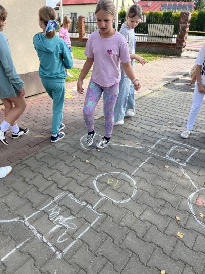 klasa czwarta ćwiczy odmianę czasownika to be grając w klasy na podwórku szkolnym