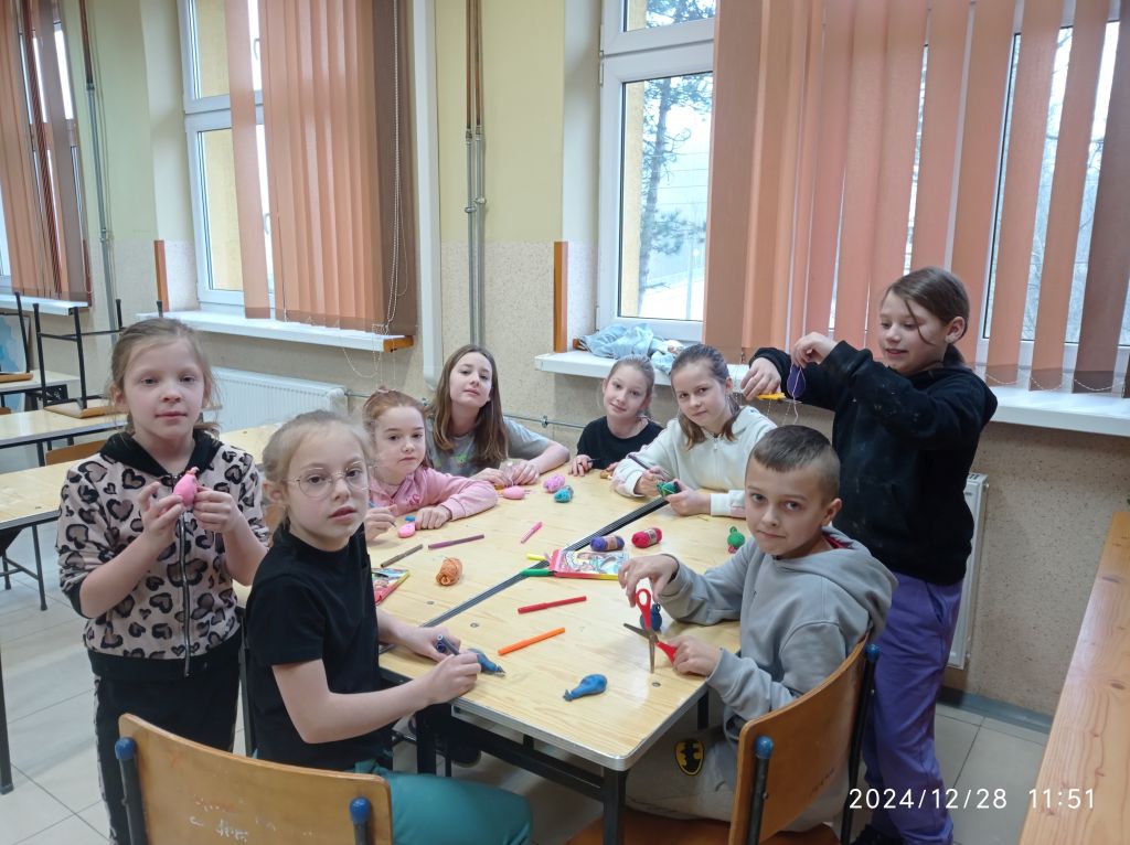 Grupa uczniów siedząc w ławkach przygotowuje prace plastyczne wykorzystując różne materiały, m.in. na ławce znajdują się balony, włóczka, mazaki, nożyczki.