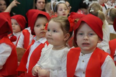 Grupka dzieci pozuje do zdjęcia. Mają białe bluzeczki, czerwone kamizelki i czerwone czapeczki na głowach