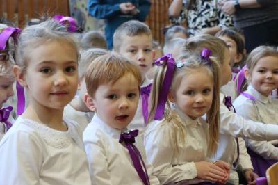 Grupka dzieci pozuje do zdjęcia. Mają białe bluzeczki, dziewczynki fioletowe kokardy we włosach.