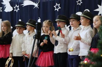 Grupka dzieci stoi w rzędzie na scenie. Śpiewają piosenkę, grają na instrumentach muzycznych.