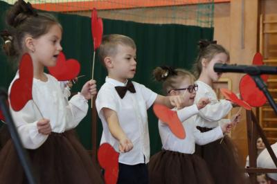 Grupka dzieci tańczy na scenie.  Machają czerwonymi sercami z papieru.