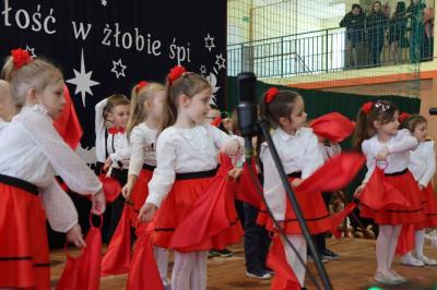 Grupka dzieci tańczy na scenie. Mają biało-czerwone ubranka, w rękach trzymają czerwone chustki.