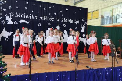 Grupka dzieci tańczy na scenie. Mają biało-czerwone ubranka, w rękach trzymają czerwone chustki.