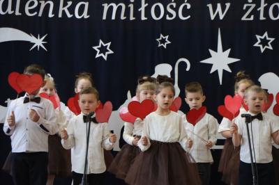 Grupka dzieci tańczy w dwóch rzędach na scenie.  Machają czerwonymi sercami z papieru