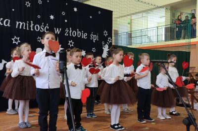 Grupka dzieci tańczy w dwóch rzędach na scenie. Mają biało-brązowe ubrania, a w rekach trzymają papierowe, czerwone serca.