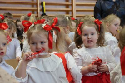 Grupka dzieci w białych bluzeczkach pozuje do zdjecia, dziewczynki mają na głowach czerwone kokardki.