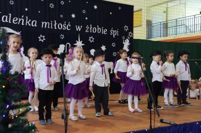 Grupka dzieci występuje w dwóch rzędach na scenie. Mają biało-fioletowe ubrania.